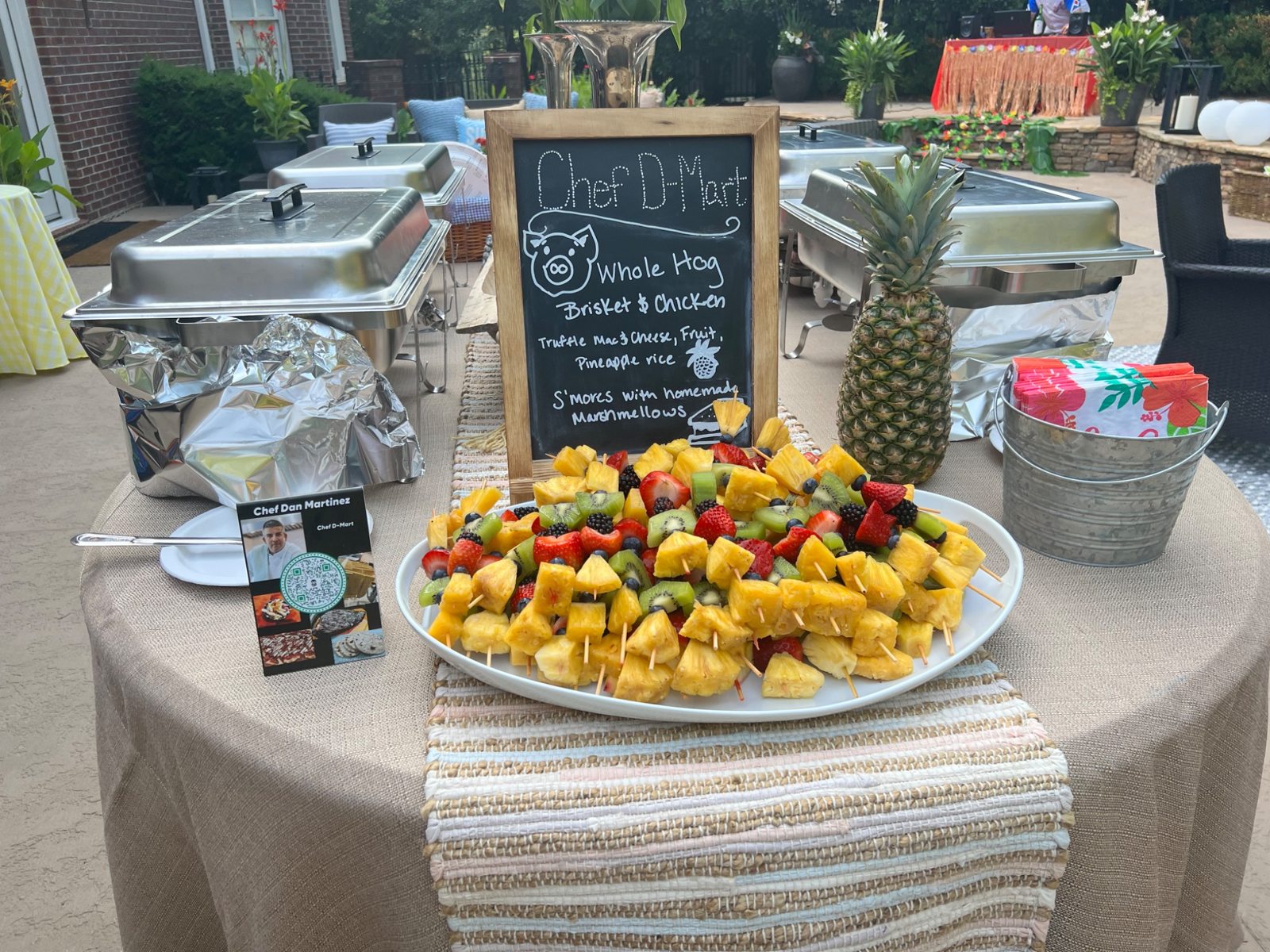 Fruit skewers, chafing dishes, and Chef D.Mart menu board at the luau buffet