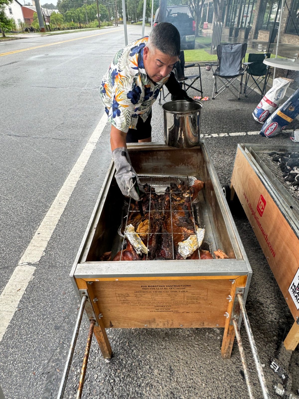 Dan tending a hog with a basting mop and a pot beside the open roasting box