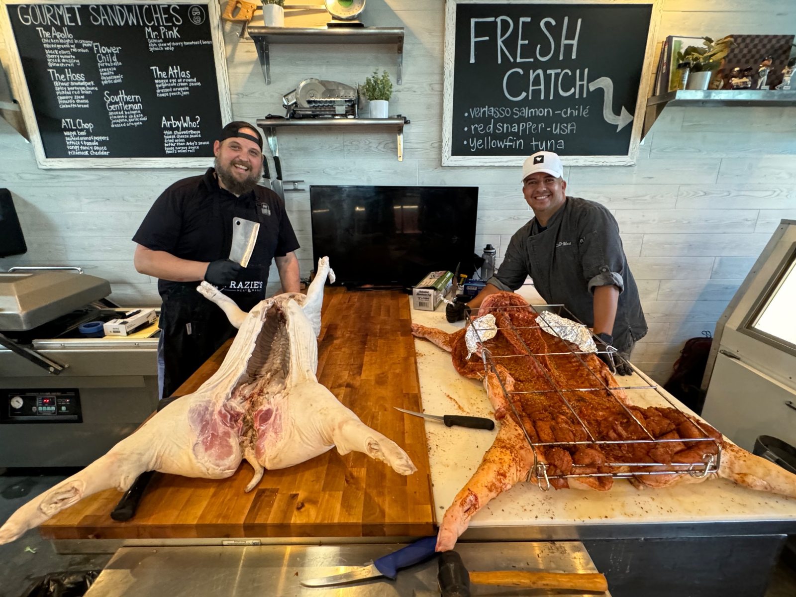 Two people standing behind two whole hogs on preparation tables at Frazie’s butcher shop